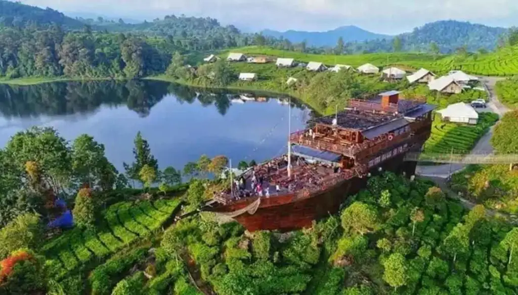 Gambar pemandangan danau di kawasan wisata Ciwidey dengan kapal besar yang berfungsi sebagai restoran terapung dan bangunan di sekitar perkebunan teh hijau.