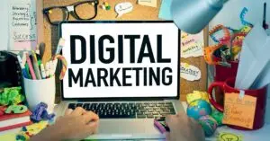 Laptop yang menampilkan teks "Digital Marketing" di tengah meja penuh alat tulis dan catatan.